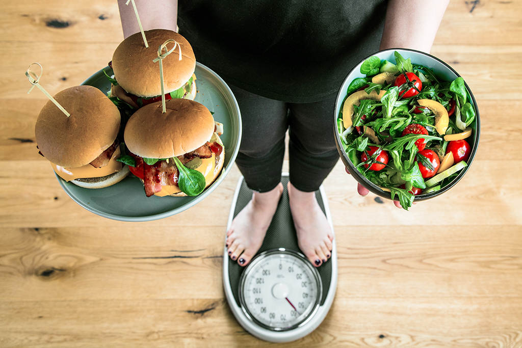 Person steht auf Waage mit Burger und Salat Person steht auf Waage mit Burger und Salat