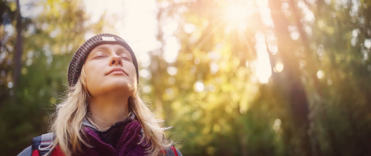 autogenes-training-kurs Eine Frau genießt mit geschlossenen Augen die Luft und den Sonnenschein im Wald.