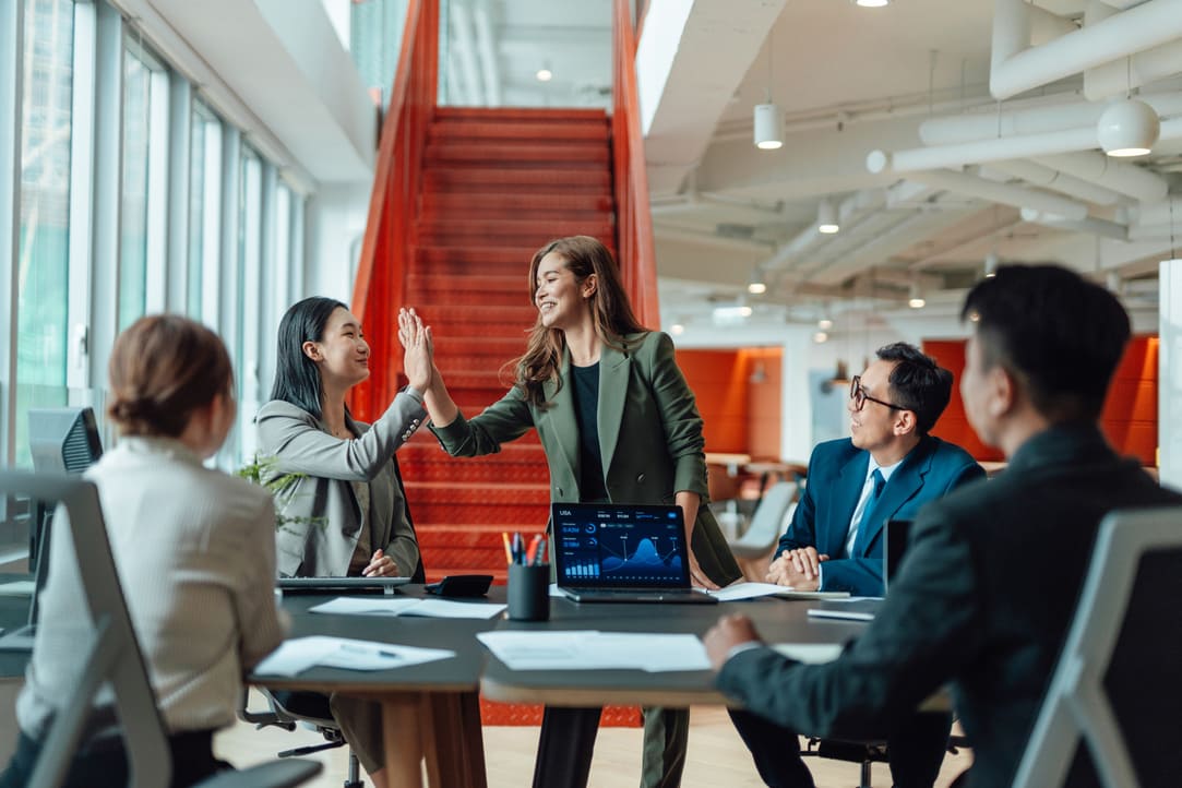 betriebliches-gesundheitsmanagement-beispiele2 Zwei Mitarbeiterinnen geben sich High-fives in einem Teammeeting