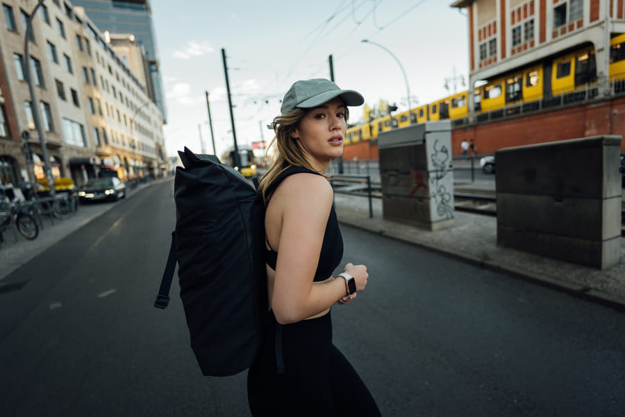 produktiver-arbeiten-artikelbild1 Eine Frau mit einer Cap und Rucksack auf überquert eine Straße und schaut in die Kamera.